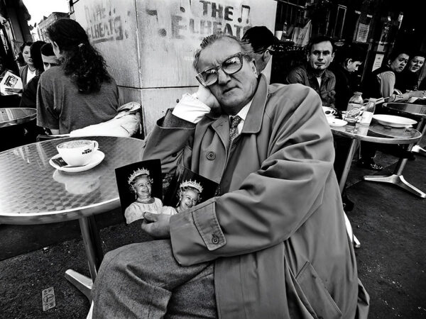 Queen of Hearts Active senior British man sitting in cafe in Soho London holding photos of Queen Elizabeth II – Fine art photography print for sale. Decoration poster for home or office wall taken by documentary travel photographer Rafael Ben Ari