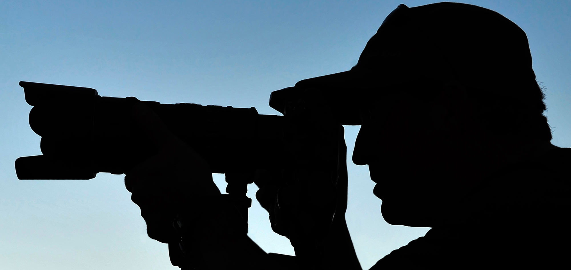 Silhouette of a photographer wearing a hat photographing with a camera and tripod nature and wildlife against blue sky.